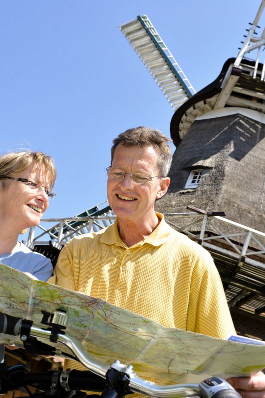 Zwei Radfahrer betrachten eine Radwanderkarte. Aus der Froschperspektive sieht man im Hintergrund die Mühle vor blauem Himmel